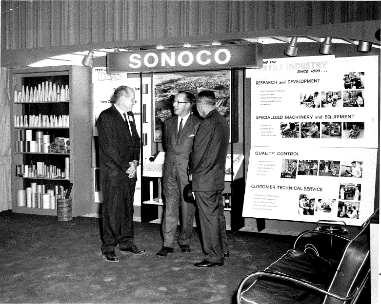 Una vieja foto en blanco y negro de tres empresarios parados frente a una pantalla SONOCO.