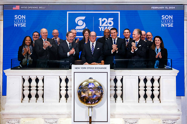 Grupo de empresarios con trajes sonriendo y aplaudiendo frente a una pared que dice "SON LISTED NYSE, SONOCO 125 Years" parada frente a la campana de la Bolsa de Valores de Nueva York.